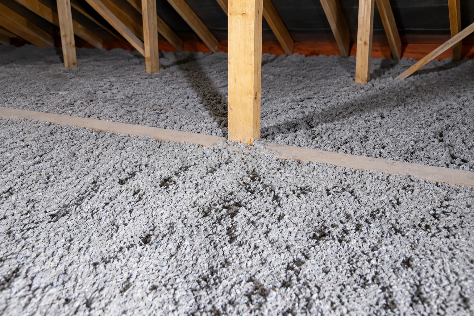 Gray blown-in insulation covers an attic floor beneath exposed wooden rafters.