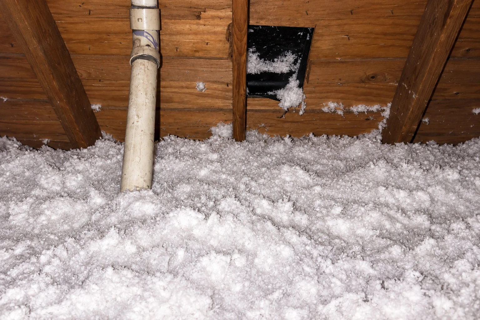 Attic floor covered in loose white blown-in insulation around a vertical pipe and roof rafters.