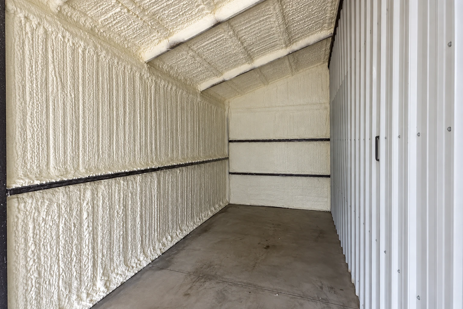 Commercial room with white spray foam insulation covering the walls and sloped ceiling beside a corrugated metal wall.