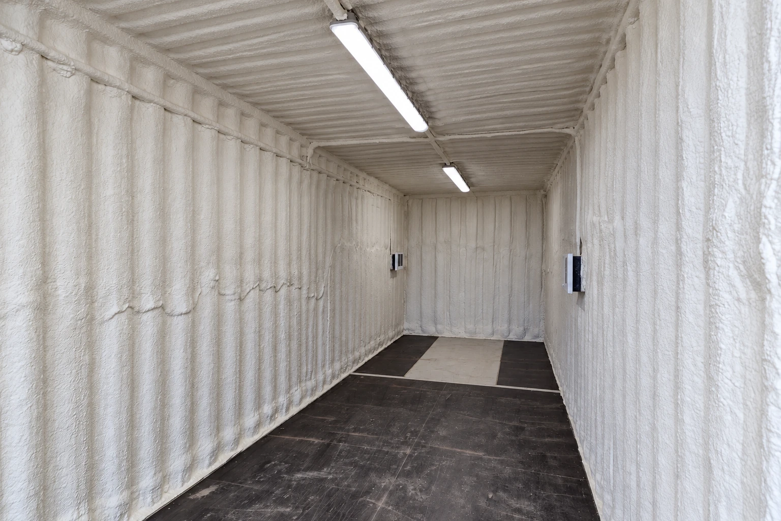 A narrow commercial interior has white spray foam insulation coating its walls and ceiling above a dark floor.