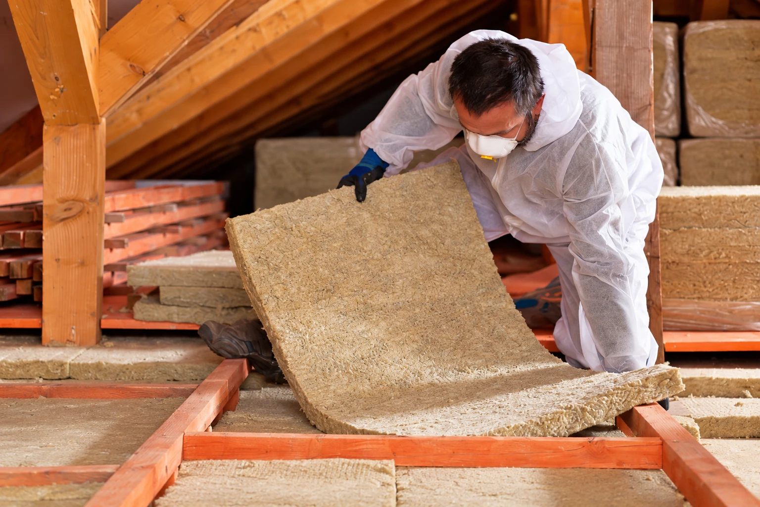 A worker in protective clothing lifts a thick tan insulation batt in an attic.